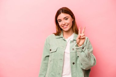 Pembe arka planda izole edilmiş genç beyaz kadın zafer işareti gösteriyor ve gülümsüyor..