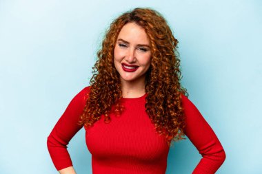 Genç, beyaz, mavi arka planda izole edilmiş, elleri kalçasında tutan, kendine güvenen bir kadın..