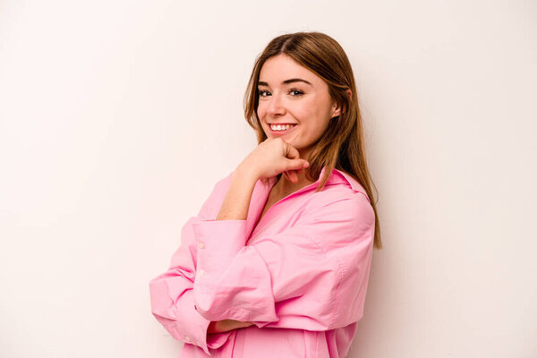 Молодая кавказская женщина на белом фоне улыбается счастливой и уверенной в себе, касаясь подбородка рукой.