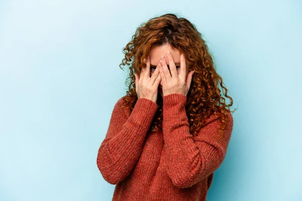 Mavi arka planda izole edilmiş genç beyaz kadın kameraya parmaklarıyla göz kırpıyor, yüzünü kapatmaktan utanıyor..