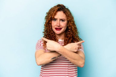 Mavi arka plan noktalarında izole edilmiş genç beyaz kadın iki seçenek arasından seçim yapmaya çalışıyor..