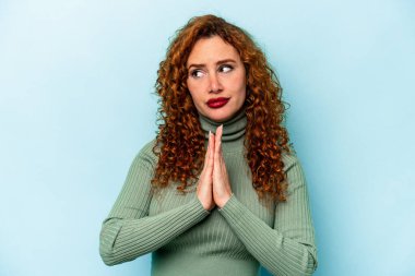 Kızıl saçlı genç bir kadın mavi arka planda izole edilmiş dua ediyor, kendini adamış, kutsal ilhamı arayan dindar biri..