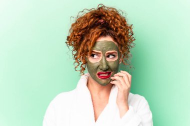 Banyodan sonra yüzünde aloe vera maskesi olan genç kızıl saçlı kadın tırnak yiyor, gergin ve endişeli..