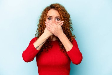 Mavi arka planda izole edilmiş kızıl saçlı genç bir kadın elleriyle ağzını kapatırken şok oldu..