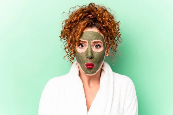 Young red hair woman with a facial aloe vera mask after a bath isolated shrugs shoulders and open eyes confused.