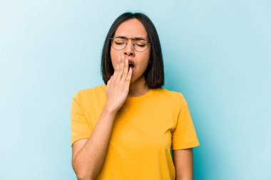 Mavi arka planda izole edilmiş genç İspanyol kadın esniyor. Ağzı eliyle kapatan yorgun bir hareket sergiliyor..