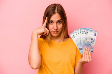 Elinde banknotlar tutan beyaz bir kadın pembe arka planda izole edilmiş, parmağıyla tapınağı işaret eden, düşünen, bir göreve odaklanmış..