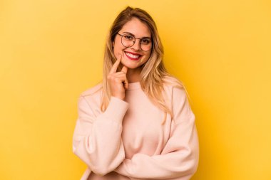 Sarı arka planda izole edilmiş genç beyaz kadın gülümsüyor ve kendinden emin, elleriyle çenesine dokunuyor..