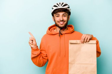 Kasklı bisiklet giyen genç İspanyol bir adam mavi arka planda izole edilmiş yiyecekleri tutuyor. Gülümsüyor ve boş alanda bir şeyler gösteriyor..