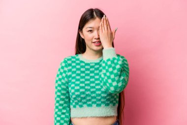 Pembe arka planda izole edilmiş genç Çinli kadın yüzünün yarısını avuç içiyle kaplayarak eğleniyor..