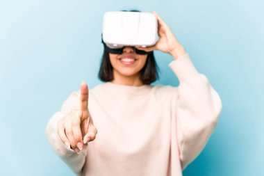 Mavi arka planda izole edilmiş sanal gözlük takan genç İspanyol kadın.