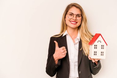 Beyaz arka planda izole edilmiş oyuncak evi tutan beyaz bir kadın gülümsüyor ve başparmağını kaldırıyor.