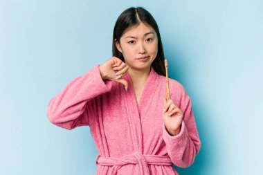 Bornoz giymiş genç Asyalı bir kadın pembe arka planda izole edilmiş dişlerini fırçalıyor ve hoş olmayan bir jest yapıyor. Anlaşmazlık konsepti.
