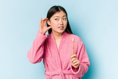 Bornoz giymiş genç Asyalı bir kadın pembe arka planda izole edilmiş dişlerini fırçalıyor bir dedikoduyu dinlemeye çalışıyor..