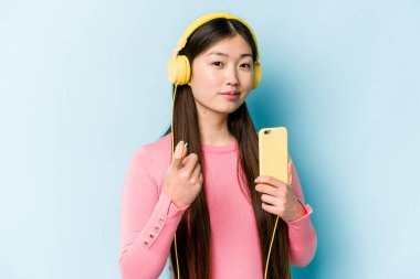 Genç Asyalı bir kadın, mavi arka planda izole edilmiş müzik dinliyor. Sanki seni davet ediyormuş gibi seni işaret ediyor..