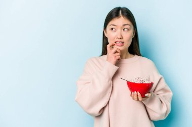 Mavi arka planda bir kase mısır gevreği tutan genç Asyalı kadın rahatlamış fotokopi odasına bakan bir şey düşünüyor..