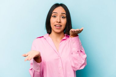 Mavi arka planda izole edilmiş genç İspanyol kadın kollarıyla ölçeği yapıyor, mutlu ve kendinden emin hissediyor..