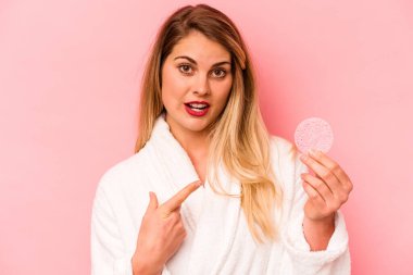 Elinde yüz süngeri tutan beyaz bir kadın var. Pembe arka planda. Yan tarafı işaret ediyor.