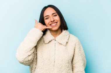 Mavi arka planda izole edilmiş genç İspanyol kadın parmaklarıyla bir cep telefonu hareketi gösteriyor..