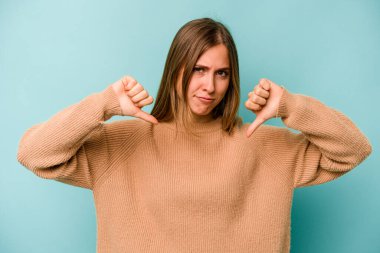 Mavi arka planda izole edilmiş genç beyaz kadın baş parmağını aşağı gösteriyor, hayal kırıklığı konsepti.
