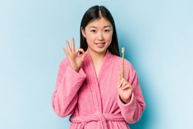 Bornoz giymiş genç Asyalı bir kadın pembe arka planda izole edilmiş dişlerini fırçalıyor neşeli ve kendinden emin bir şekilde iyi niyet gösteriyor..