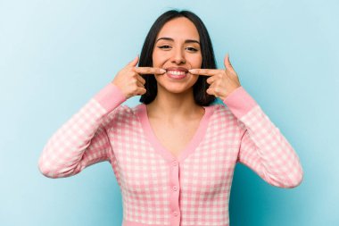 Mavi arka planda izole edilmiş genç İspanyol kadın gülümsüyor, parmaklarını ağıza doğrultuyor..