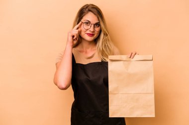 Restoran garsonu Kafkasyalı kadın bej arka planda izole edilmiş bir çanta tutuyor. Şakağı parmağıyla işaret ediyor, düşünüyor ve bir göreve odaklanıyor..