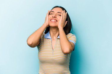 Mavi arka planda izole edilmiş genç İspanyol kadın mutlu bir şekilde ellerini başının üstünde tutarak gülüyor. Mutluluk kavramı.