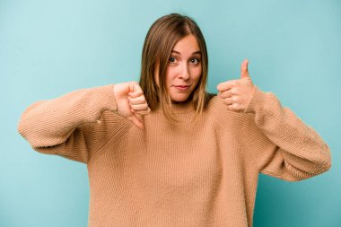 Mavi arka planda izole edilmiş genç beyaz kadın baş parmaklarını aşağı ve yukarı kaldırıyor, zor seçim konsepti