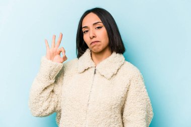Mavi arka planda izole edilmiş genç İspanyol kadın bir göz kırptı ve elleriyle iyi bir jest yaptı..