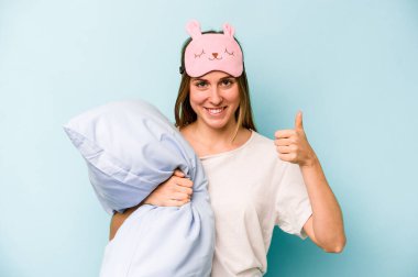 Mavi arka planda izole edilmiş pijama giyen beyaz bir kadın gülümsüyor ve başparmağını kaldırıyor.