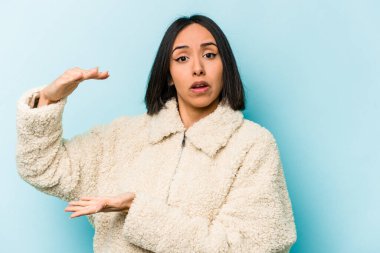 Mavi arka planda izole edilmiş genç İspanyol kadın iki eliyle bir şey tutuyor, ürün sunumu..