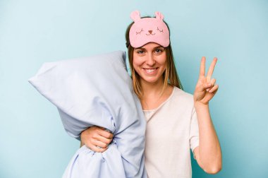 Mavi arka planda izole edilmiş pijama giyen beyaz bir kadın iki numarayı parmaklarıyla gösteriyor..