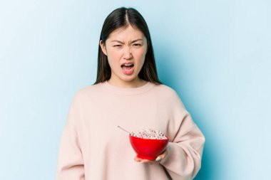 Genç Asyalı bir kadın elinde bir kase mısır gevreği tutarken mavi arka planda çok sinirli ve agresif çığlıklar atıyor..