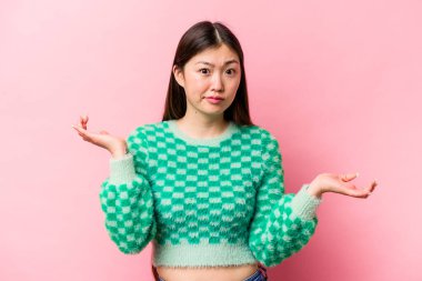 Genç Çinli bir kadın pembe arka planda izole edilmiş şüphe içinde ve sorgulama sırasında omuz silkiyor..