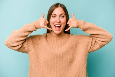 Mavi arka planda izole edilmiş genç beyaz kadın parmaklarıyla bir cep telefonu hareketi gösteriyor..