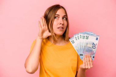 Elinde banknotlar tutan beyaz bir kadın pembe arka planda izole edilmiş bir şekilde bir dedikoduyu dinlemeye çalışıyor..