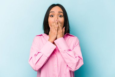 Mavi arka planda izole edilmiş genç İspanyol kadın elleriyle ağzını kapatırken şok oldu..