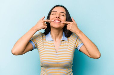 Mavi arka planda izole edilmiş genç İspanyol kadın gülümsüyor, parmaklarını ağıza doğrultuyor..