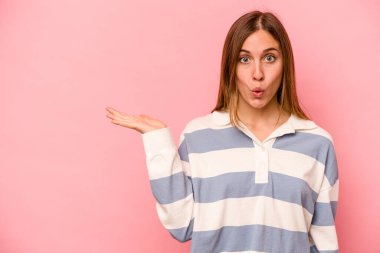 Pembe arka planda izole edilmiş genç beyaz kadın, avuç içi fotokopi alanını elinde tutmaktan etkilenmiş..