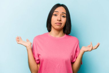 Latin kökenli genç bir kadın, mavi arka planda izole edilmiş. Sorgulama sırasında omuz silkiyor ve şüphe duyuyor..