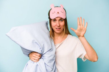 Mavi arka planda izole edilmiş pijama giyen beyaz bir kadın gülümseyerek beş numarayı parmaklarıyla gösteriyor..