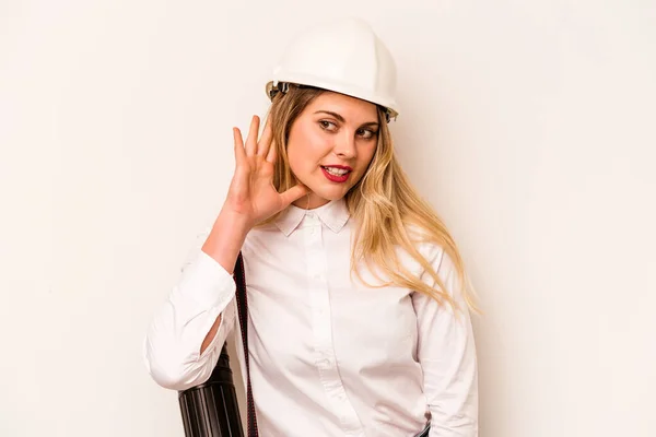 Young architect woman with helmet and holding blueprints isolated on white background trying to listening a gossip.