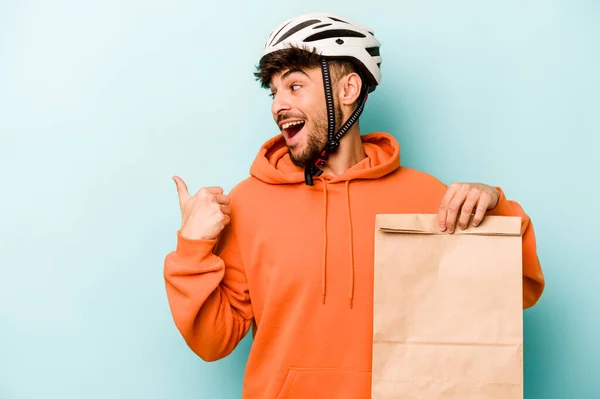 Young hispanic man wearing a helmet bike holding a take away food isolated on blue background points with thumb finger away, laughing and carefree.