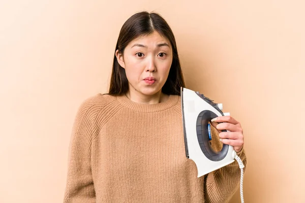 Young Asian Woman Holding Iron Isolated Beige Background Shrugs Shoulders — Stock Photo, Image