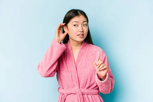 Young asian woman holding razor blade isolated on blue background trying to listening a gossip.