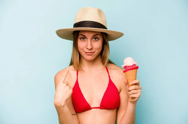 年轻的高加索女人穿着比基尼 拿着一只蓝色背景的冰淇淋 用手指指着你 好像邀请你走近些 — 图库照片