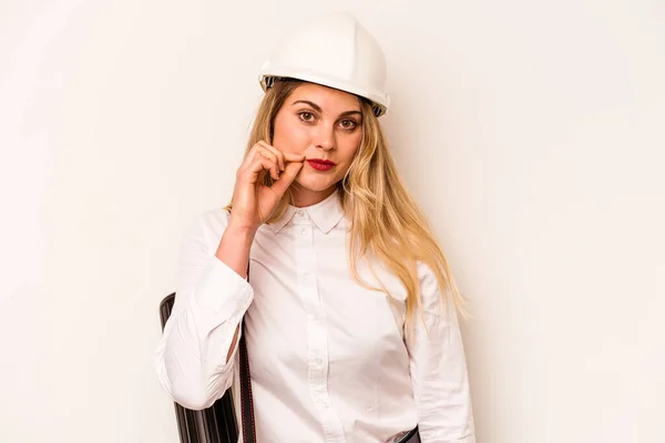 Young architect woman with helmet and holding blueprints isolated on white background with fingers on lips keeping a secret.