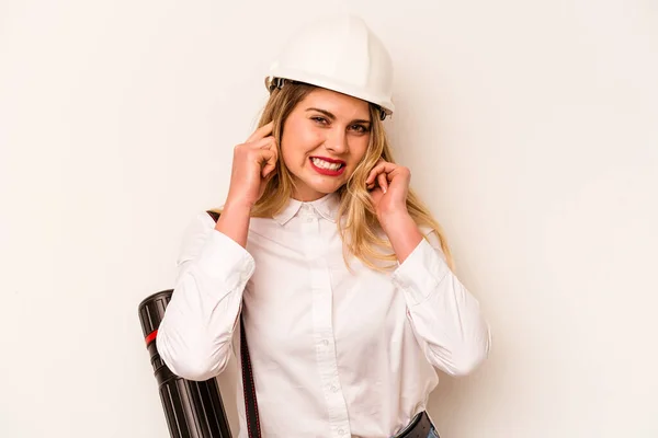 Young architect woman with helmet and holding blueprints isolated on white background covering ears with hands.