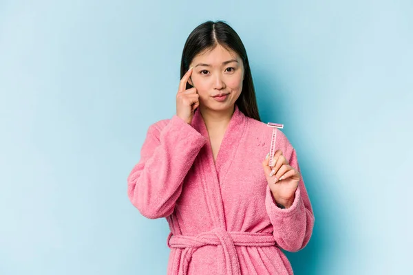 年轻的亚洲女人拿着剃须刀刀片 隔离在蓝色的背景下 用手指 专注于一项任务 指向寺庙 — 图库照片